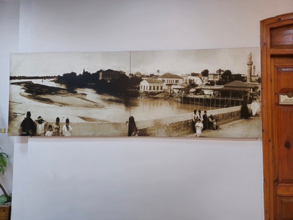 Tarihi bir fotoğraf, nehir kenarında yürüyen insan grubu ve çevresindeki binalarla birlikte panoramik bir manzara sunuyor.